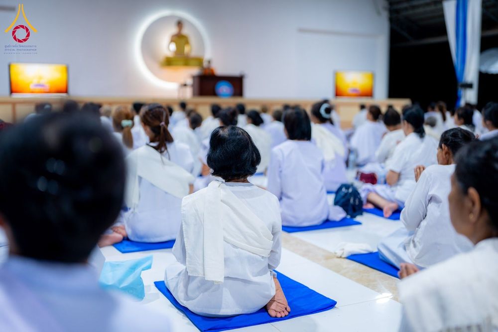กลุ่มผู้เข้าร่วมโครงการอบรมสวมชุดสีขาวนั่งปฏิบัติธรรมบนเบาะสีน้ำเงินภายในอาคาร