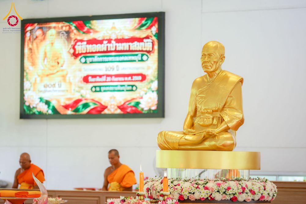 รูปหล่อพระภิกษุสีทองตั้งอยู่หน้าจอแสดงภาพป้ายประชาสัมพันธ์งานบุญและพระภิกษุสงฆ์