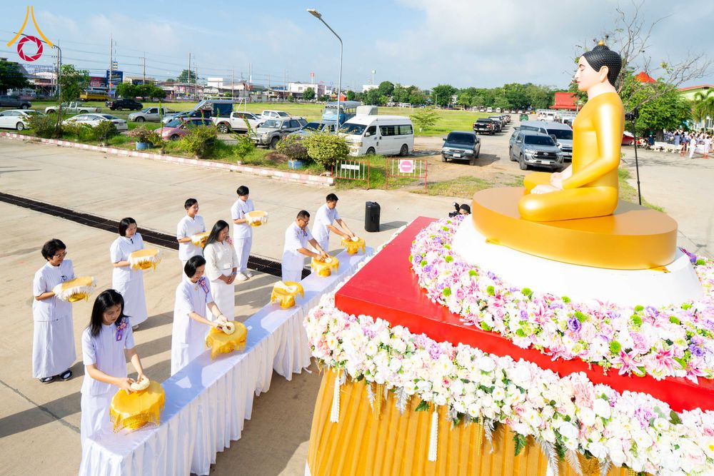 กลุ่มคนชุดขาวกำลังประกอบพิธีทางศาสนาหน้าองค์พระพุทธรูปสีทองที่ประดับด้วยดอกไม้หลากสี