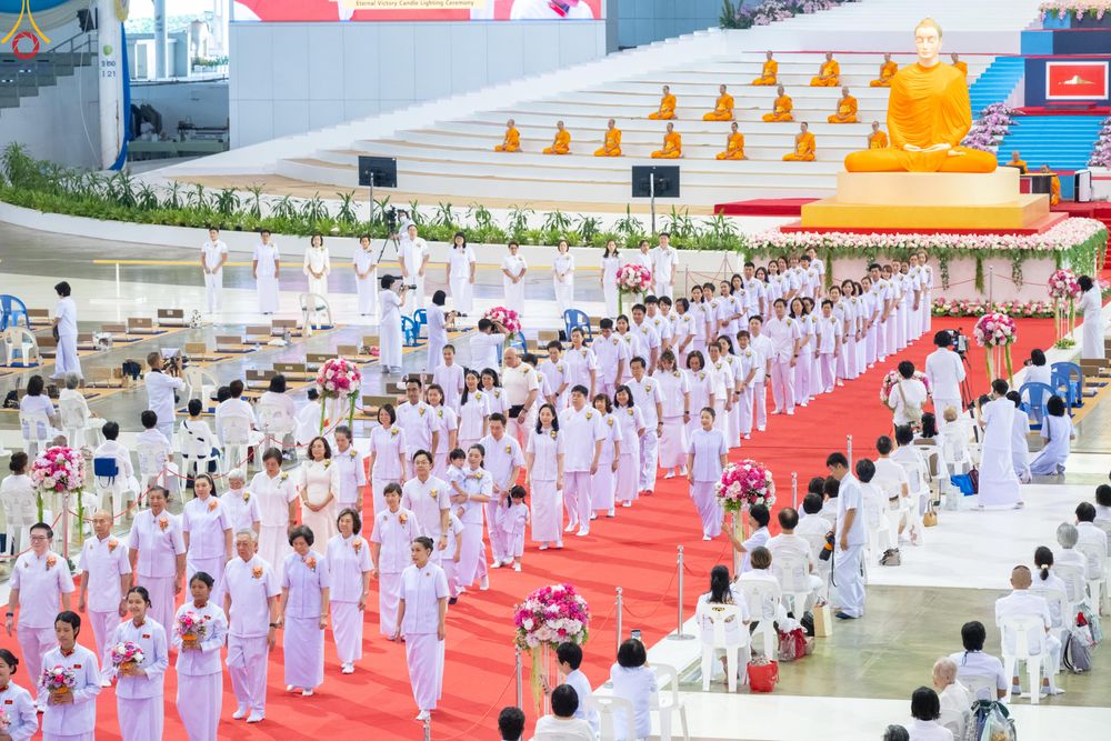 กลุ่มคนสวมชุดขาวเดินแถวบนพรมแดงในพิธีทางศาสนา โดยมีพระภิกษุสงฆ์และพระพุทธรูปเป็นฉากหลัง