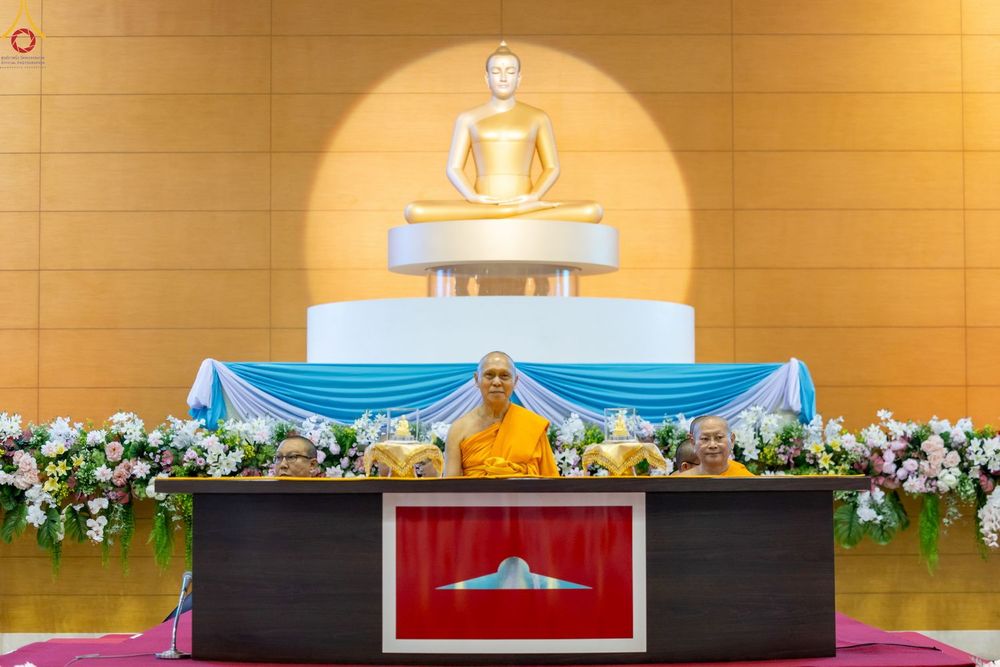 พระภิกษุสงฆ์นั่งบนเวทีหน้าพระพุทธรูปในห้องโถงที่มีการตกแต่งด้วยดอกไม้