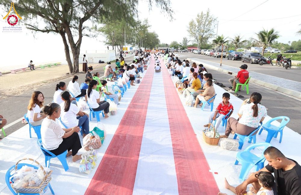 ประชาชนนั่งรอตักบาตรพระสงฆ์ริมชายหาดโดยมีผ้าปูสีขาวแดงวางยาวอยู่ตรงกลาง