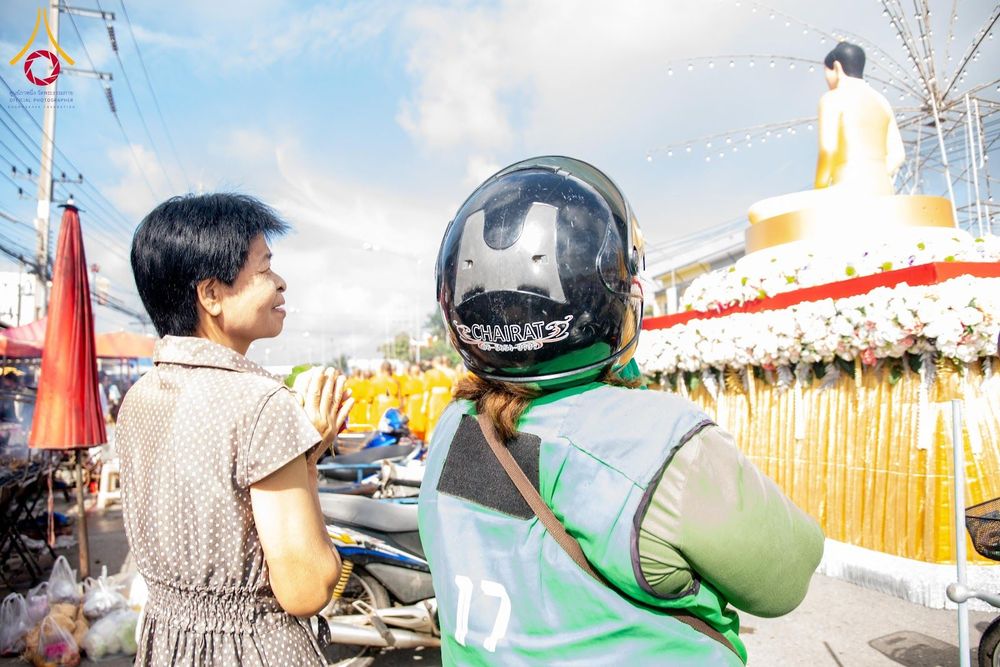 หญิงไทยพนมมือไหว้พระพุทธรูป โดยมีผู้สวมหมวกกันน็อกและเสื้อกั๊กสีเขียวยืนอยู่ข้างๆ ในบรรยากาศงานพิธีทางศาสนา