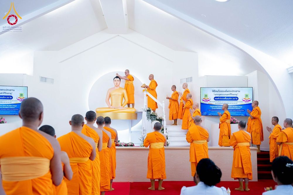 พระภิกษุสงฆ์ในจีวรสีส้มกำลังประกอบพิธีบนแท่นประดิษฐานพระประธานสีทองภายในห้องโถงสีขาวที่มีผู้เข้าร่วมพิธีจำนวนมาก