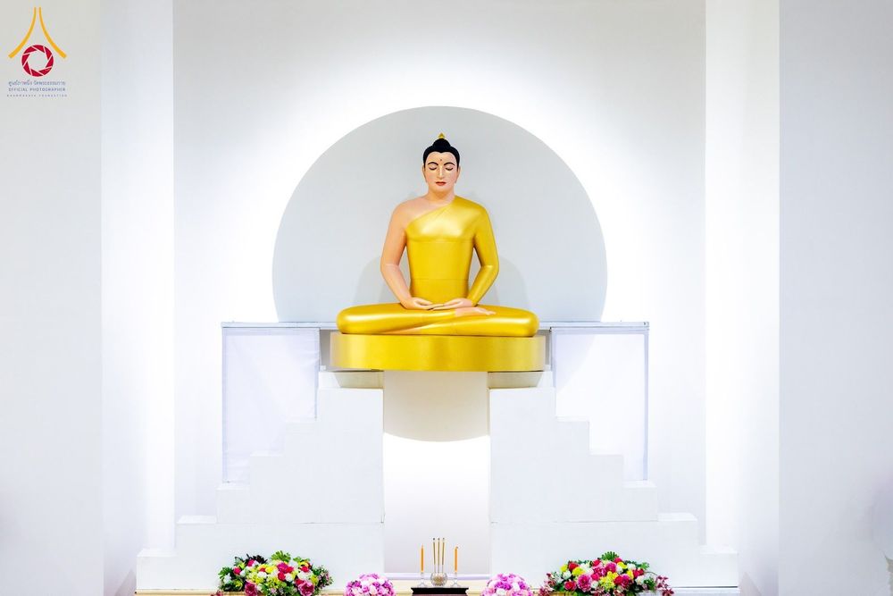 พระพุทธรูปสีทองปางสมาธิประดิษฐานบนฐานสีขาว มีฉากหลังเป็นวงกลมสีขาวและประดับด้วยดอกไม้สด