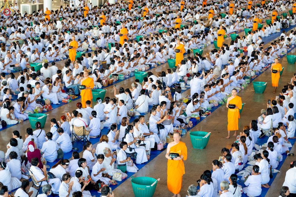 พุทธศาสนิกชนชุดขาวจำนวนมากนั่งเรียงแถวรอตักบาตรพระภิกษุในอาคารกว้างขวาง