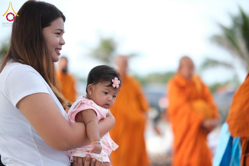 หญิงสาวอุ้มทารกน้อยในอ้อมแขนขณะร่วมพิธีตักบาตรที่มีพระสงฆ์เดินผ่าน