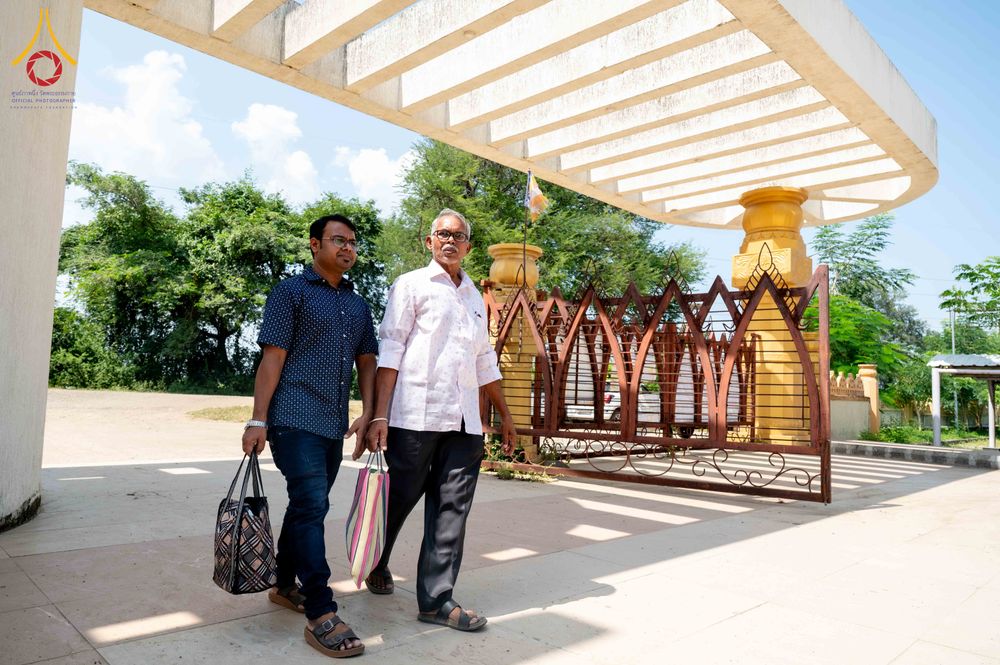 ชายสองคนกำลังเดินผ่านซุ้มประตูทางเข้าที่มีโครงสร้างหลังคาคอนกรีตและรั้วเหล็กดัดสีน้ำตาล