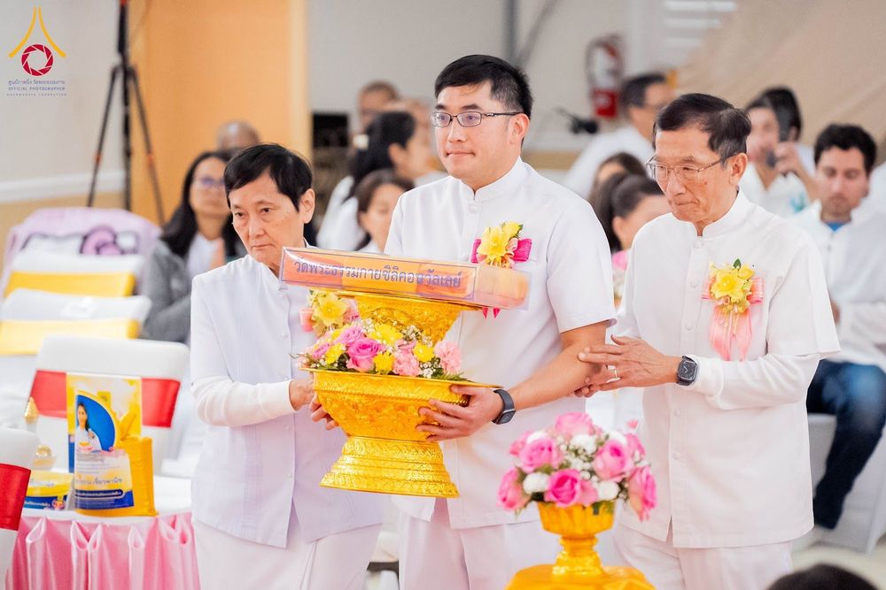 กลุ่มคนในชุดสีขาวกำลังถือพานพุ่มดอกไม้และผ้าไตรในพิธีทางศาสนา
