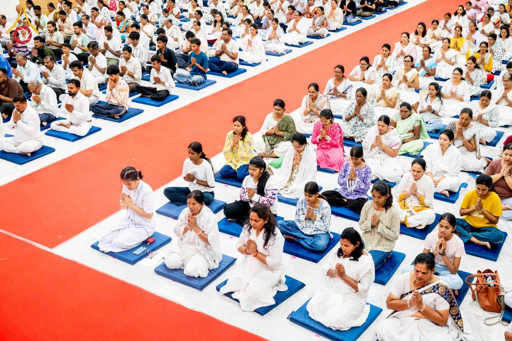 กลุ่มผู้ปฏิบัติธรรมสวมชุดสีขาวนั่งสมาธิบนเบาะรองนั่งภายในอาคารที่มีทางเดินปูพรมสีแดง
