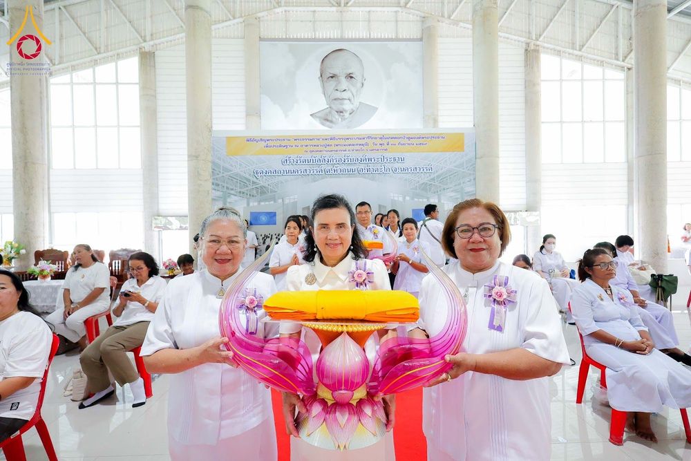 กลุ่มผู้หญิงในชุดขาวถือพานพุ่มผ้าไตรในพิธีทางศาสนาภายในอาคารที่มีฉากหลังเป็นภาพพระสงฆ์