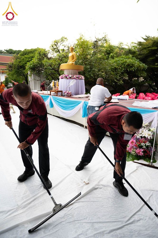ชายสองคนกำลังใช้ไม้รีดน้ำทำความสะอาดพื้นพลาสติกสีขาวในบริเวณสถานที่จัดงาน