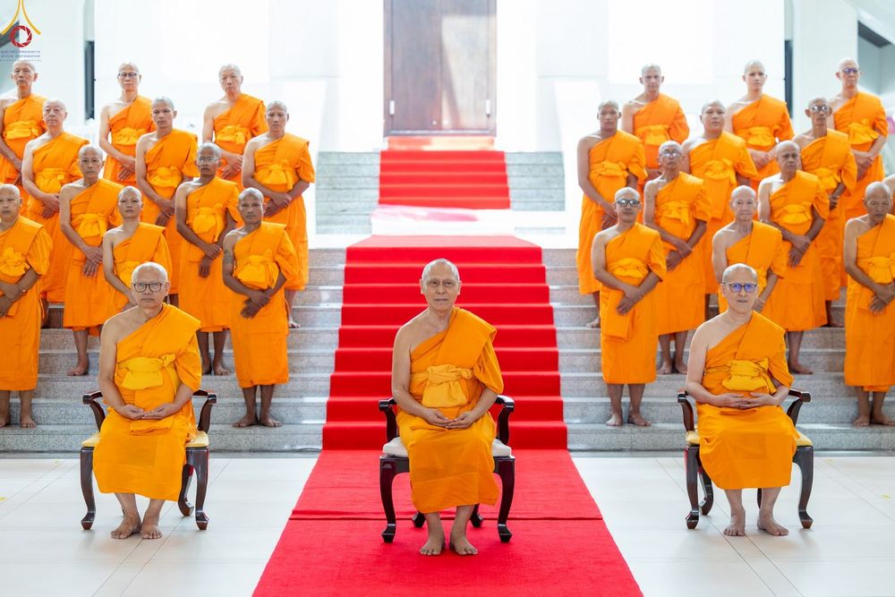 กลุ่มพระภิกษุและนาคยืนเรียงแถวบนบันไดหน้าโบสถ์ที่มีพรมแดงปูพื้น ในพิธีบรรพชาอุปสมบท