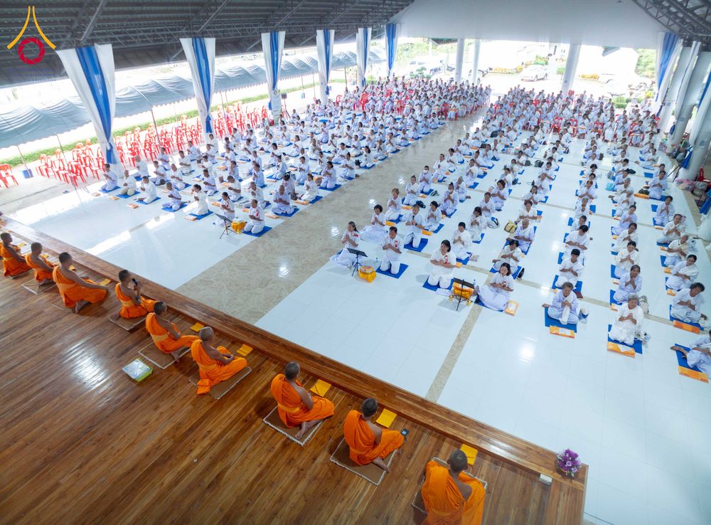 กลุ่มผู้ปฏิบัติธรรมชุดขาวนั่งเรียงแถวบนพื้นอาคารและพระภิกษุชุดจีวรสีส้มบนเวทีไม้