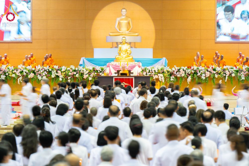 พุทธศาสนิกชนสวมชุดขาวนั่งร่วมพิธีทอดผ้าป่าสมทบกฐินภายในสภาธรรมกายสากลที่มีพระภิกษุสงฆ์และพระพุทธรูปบนเวที