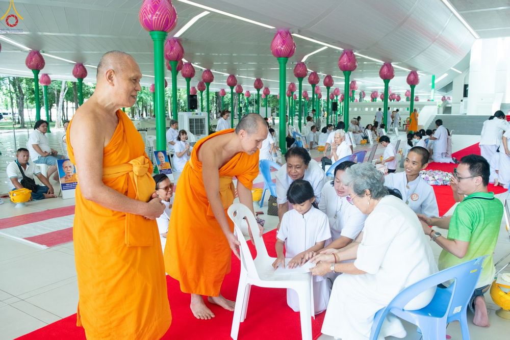 พระภิกษุสงฆ์และพุทธศาสนิกชนในชุดขาวกำลังทำกิจกรรมร่วมกันภายในอาคารที่มีเสารูปดอกบัว