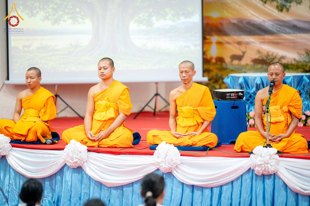 พระสงฆ์สี่รูปนั่งสมาธิบนเวทีที่มีการตกแต่งด้วยผ้าสีฟ้าและขาว โดยมีพระสงฆ์รูปหนึ่งกำลังใช้ไมโครโฟน