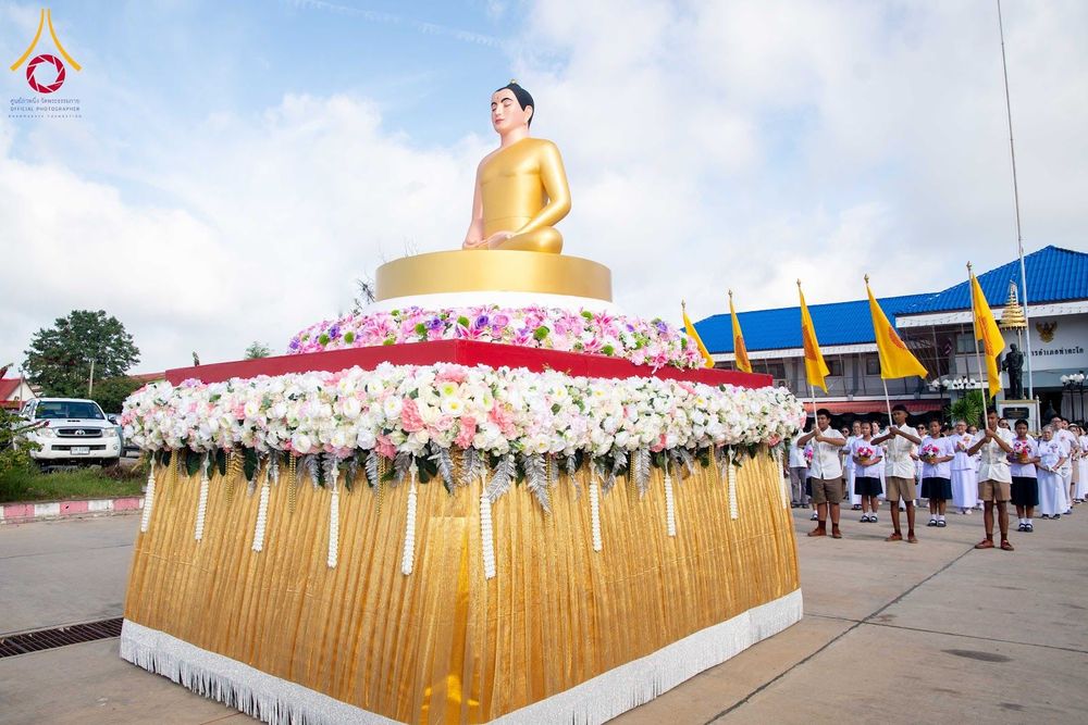 พระพุทธรูปสีทองบนฐานประดับดอกไม้และกลุ่มคนถือธงในพิธีทางศาสนา