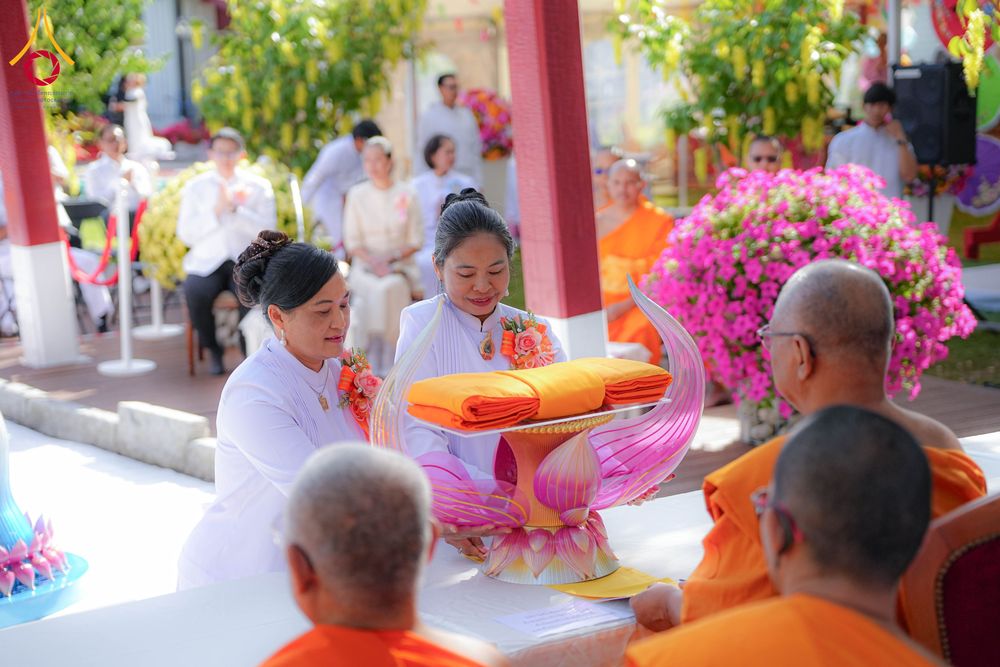 หญิงสองคนในชุดขาวกำลังประคองพานผ้าไตรจีวรเพื่อถวายแด่พระภิกษุสงฆ์ในพิธีทำบุญ