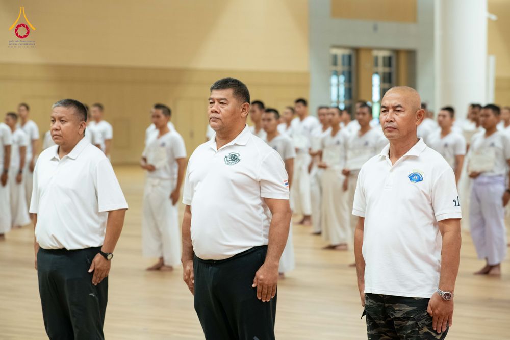 กลุ่มชายในชุดเสื้อโปโลสีขาวและชุดปฏิบัติธรรมสีขาว ยืนเรียงแถวอย่างเป็นระเบียบภายในอาคาร