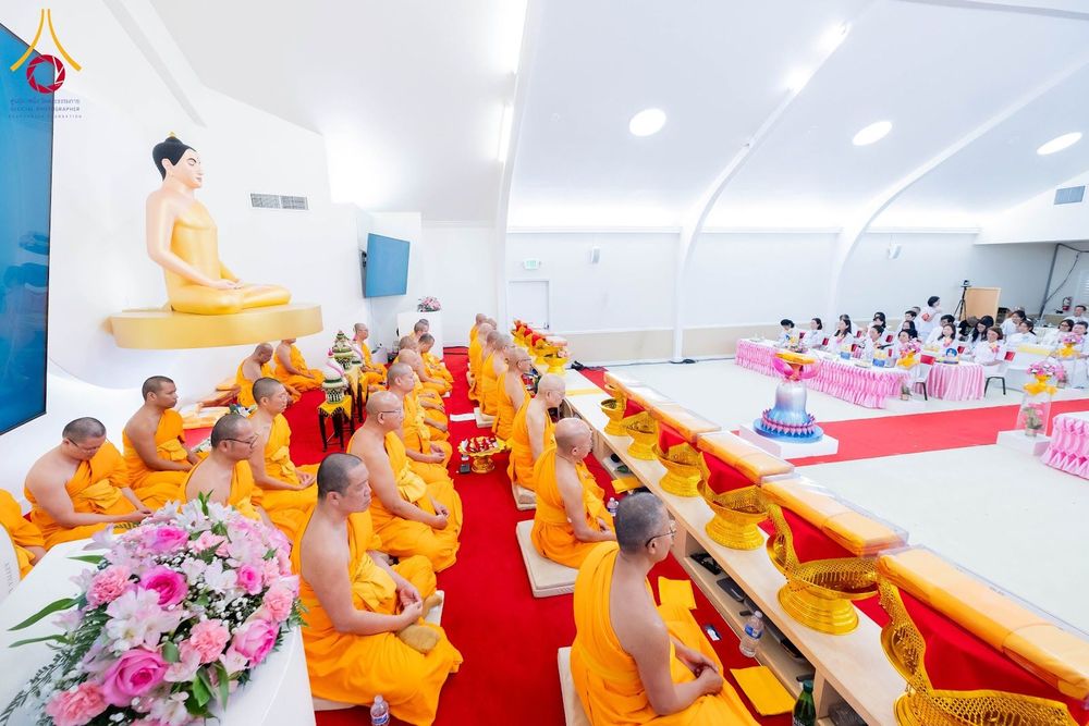 พระภิกษุสงฆ์ห่มจีวรสีเหลืองทองนั่งเรียงแถวประกอบพิธีทางศาสนาภายในอาคารที่มีพระพุทธรูปประดิษฐานอยู่ด้านหน้า