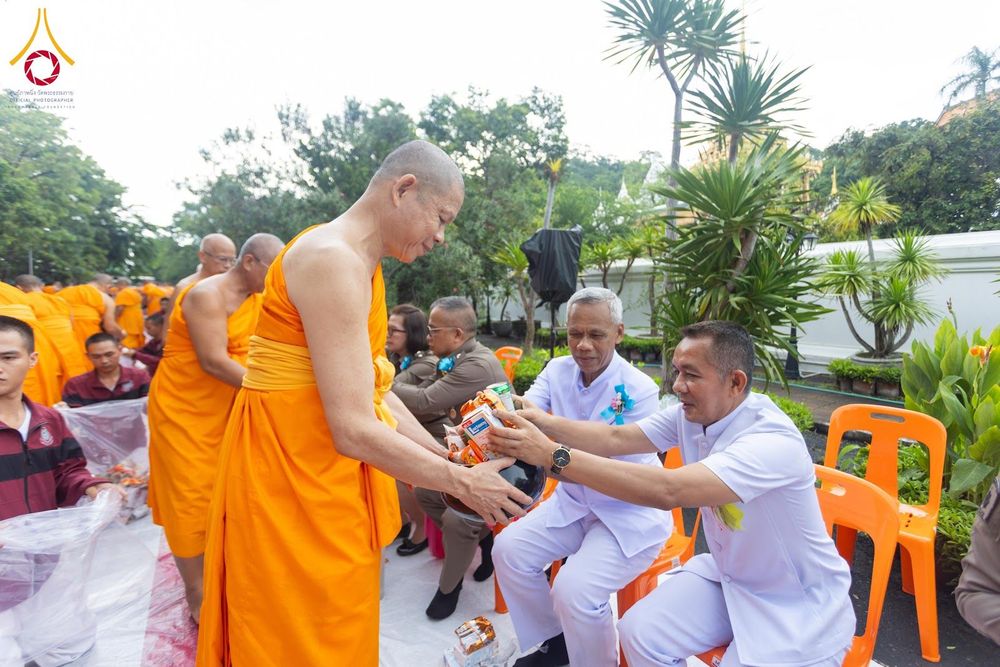 ชายในชุดขาวกำลังประเคนสิ่งของใส่บาตรให้แก่สามเณรในพิธีตักบาตร