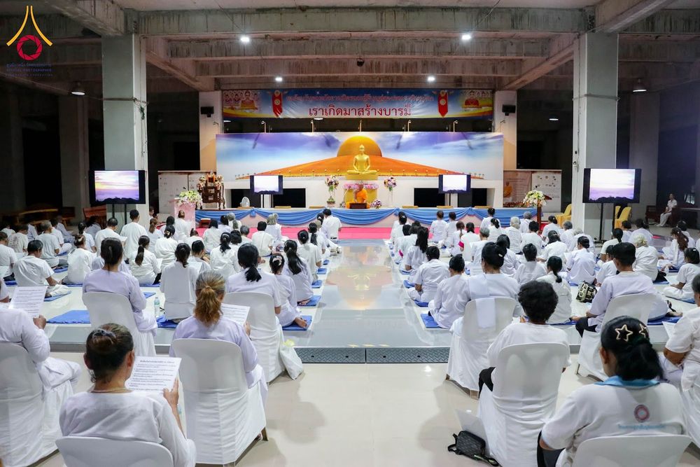 ผู้เข้าร่วมอบรมในชุดสีขาวนั่งเรียงแถวฟังธรรมภายในหอประชุมที่มีพระพุทธรูปประดิษฐานอยู่ด้านหน้า