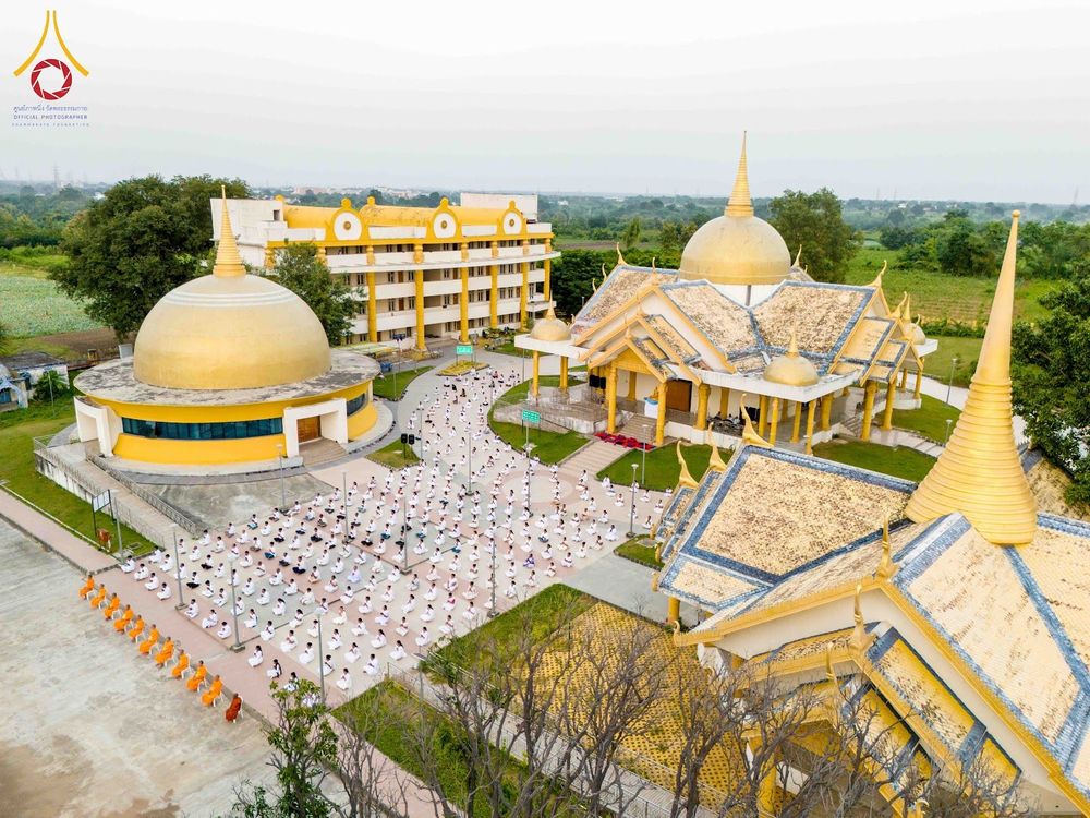 กลุ่มผู้ปฏิบัติธรรมชุดขาวนั่งสมาธิในลานกว้างหน้าอาคารสถาปัตยกรรมสีเหลืองทอง