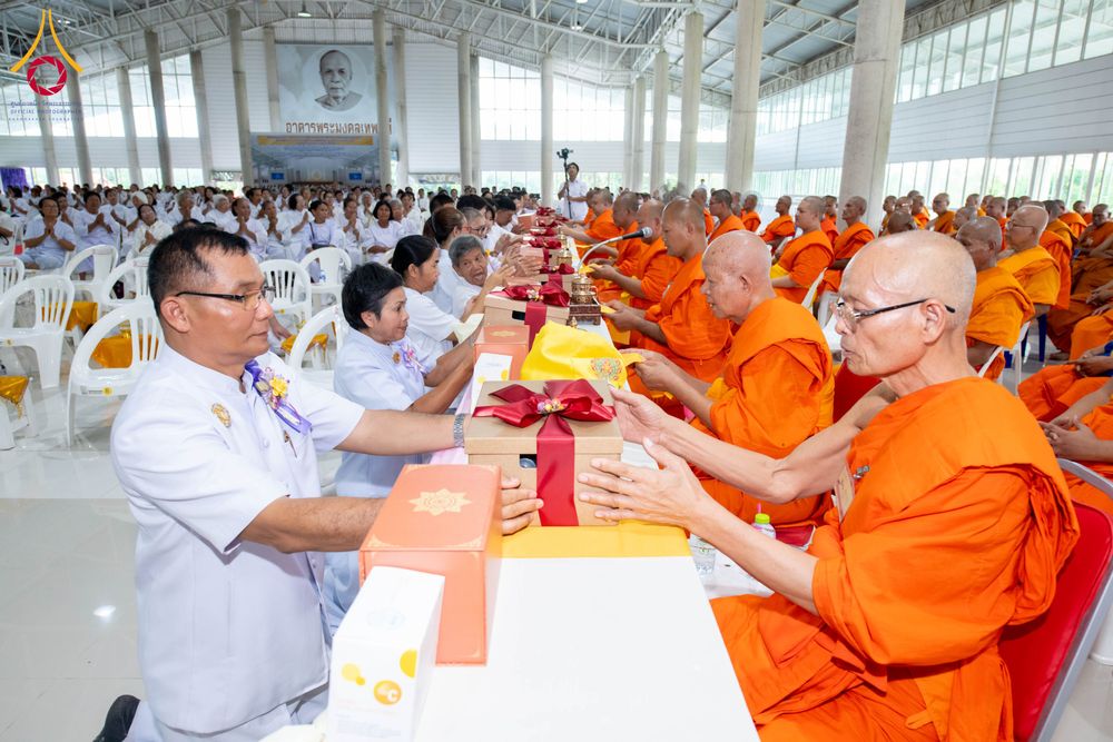 ชายในชุดขาวกำลังถวายกล่องของขวัญผูกโบว์สีแดงแด่พระภิกษุสงฆ์ภายในอาคารที่มีสาธุชนนั่งร่วมพิธีจำนวนมาก