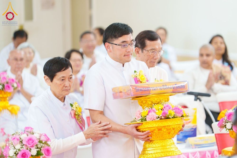 กลุ่มพุทธศาสนิกชนในชุดสีขาวกำลังถือพานพุ่มดอกไม้และผ้าไตรในพิธีทอดผ้าป่า