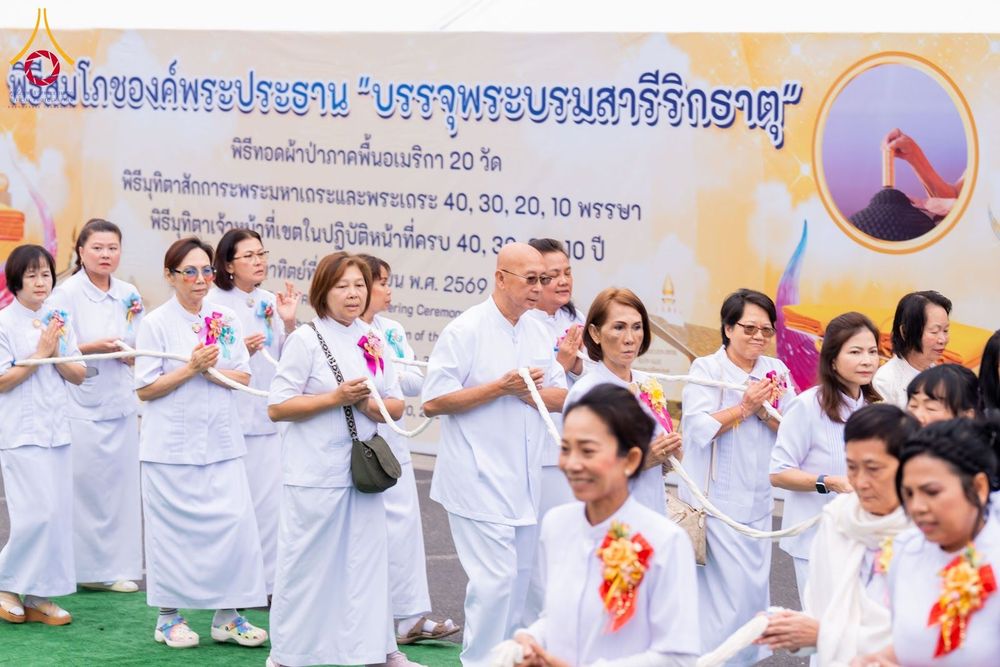 กลุ่มคนสวมชุดสีขาวถือสายสิญจน์ร่วมกันในพิธีทางศาสนาหน้าป้ายไวนิล