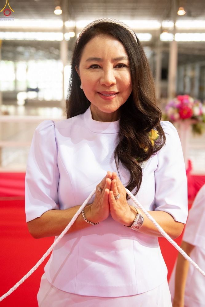 สุภาพสตรีสวมชุดสีขาวพนมมือยิ้มแย้ม มีสายสิญจน์พาดผ่านด้านหน้า