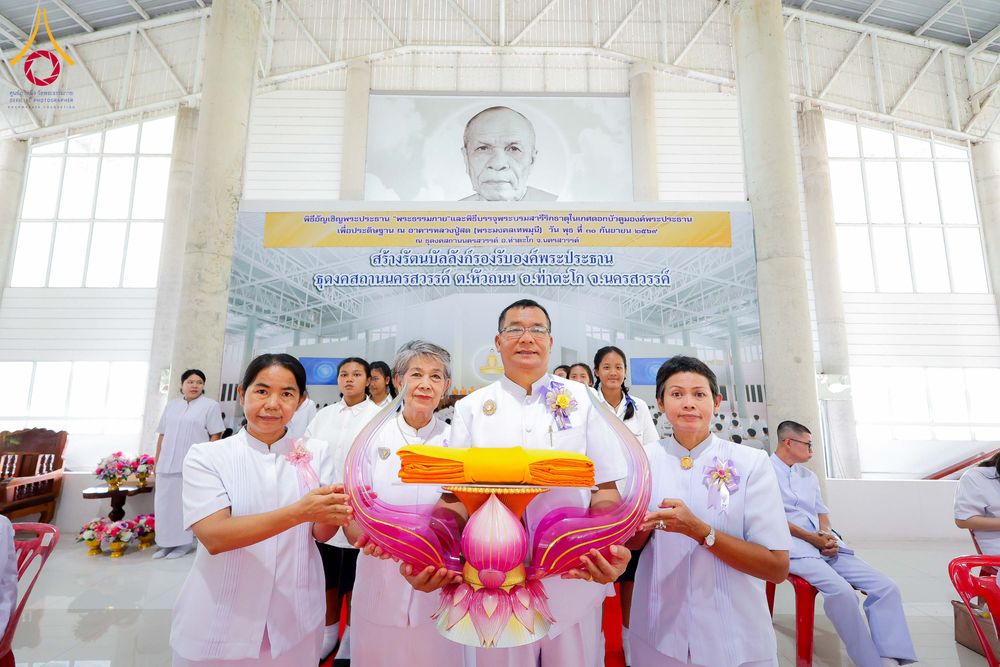 กลุ่มคนในชุดขาวกำลังถือพานพุ่มดอกบัวสีชมพูที่มีผ้าไตรวางอยู่ด้านบนภายในอาคารที่มีฉากหลังเป็นป้ายพิธีและภาพพระมงคลเทพมุนี