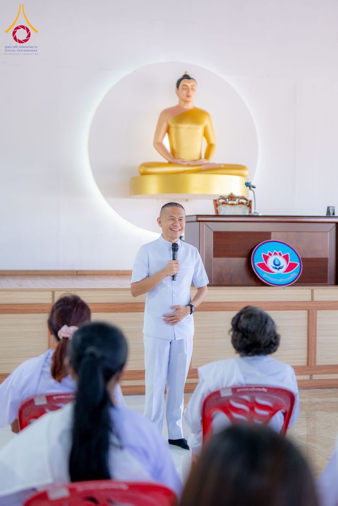 วิทยากรชายสวมชุดสีขาวกำลังยืนบรรยายธรรมต่อหน้าผู้เข้าร่วมโครงการอบรมที่มีพระพุทธรูปสีทองเป็นฉากหลัง