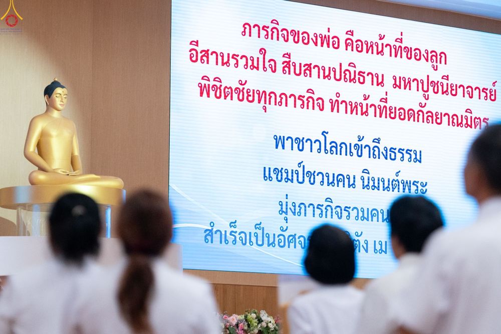 กลุ่มคนสวมชุดขาวหันหลังให้กล้องมองไปยังจอโปรเจกเตอร์ที่มีข้อความภาษาไทยและพระพุทธรูปสีทอง