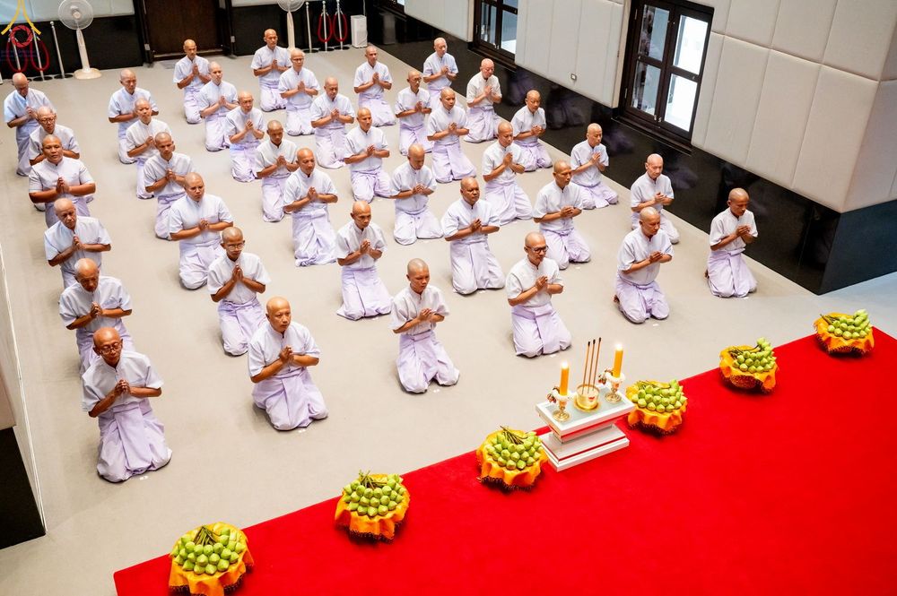 กลุ่มผู้เข้าร่วมพิธีบรรพชาอุปสมบทในชุดสีขาวนั่งพนมมือเรียงแถวบนพื้นภายในอาคาร