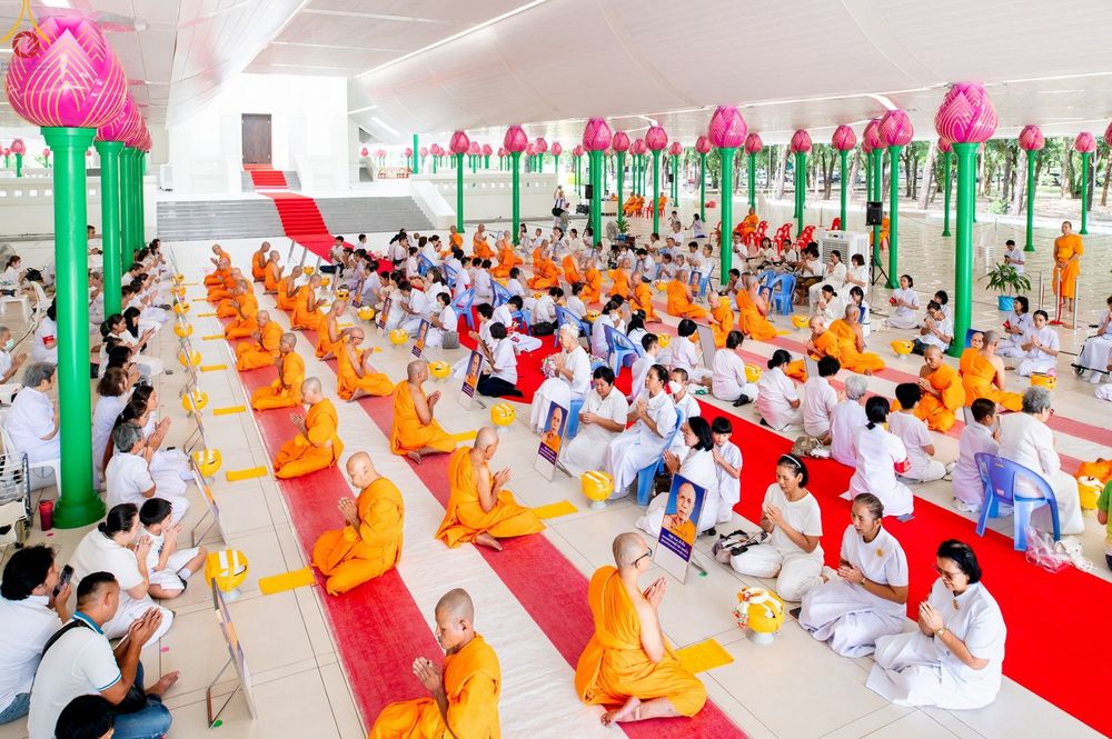 พระภิกษุ สามเณร และพุทธศาสนิกชนชุดขาวนั่งพนมมือร่วมพิธีบรรพชาอุปสมบทภายในอาคารที่มีเสารูปดอกบัวประดับ