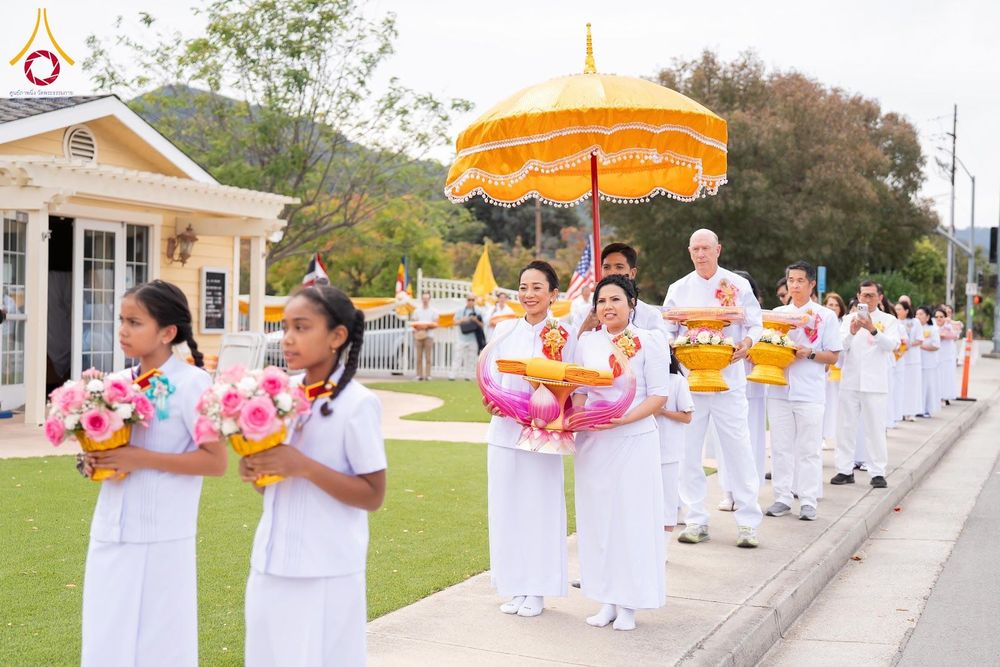 กลุ่มคนในชุดขาวเดินแถวถือพานดอกไม้และผ้าไตรในพิธีทางศาสนาพุทธ