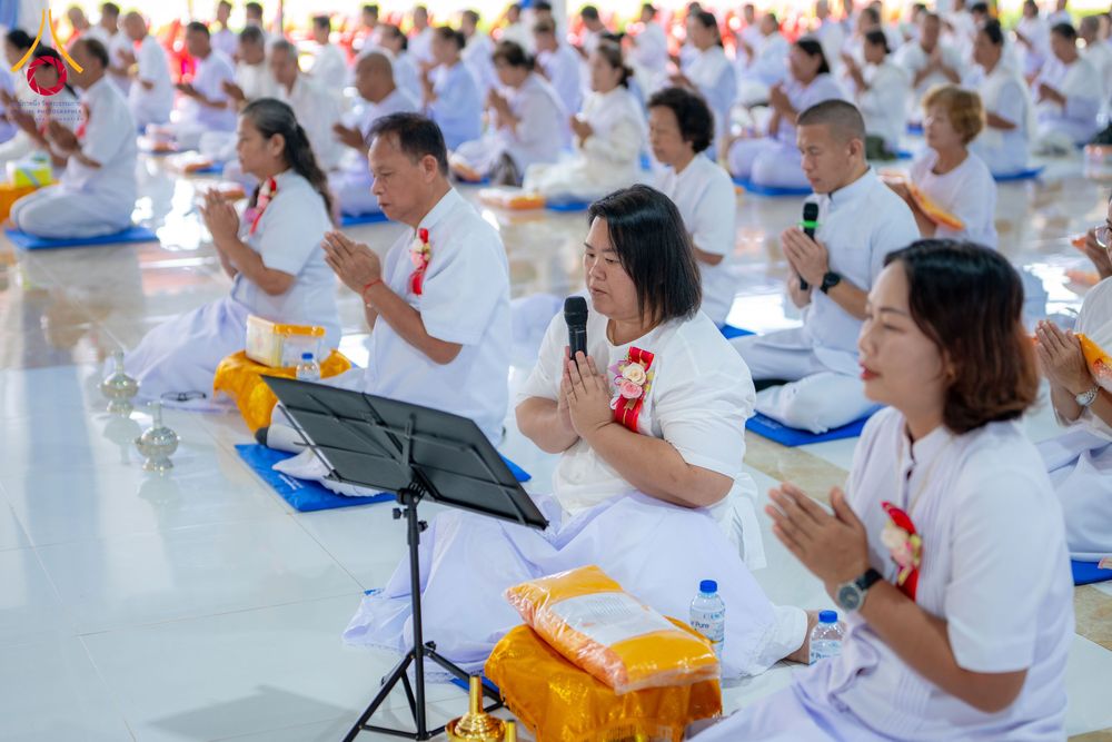 กลุ่มคนในชุดสีขาวนั่งพนมมือปฏิบัติธรรมภายในอาคาร โดยมีผู้หญิงถือไมโครโฟนนำสวดมนต์อยู่ด้านหน้า