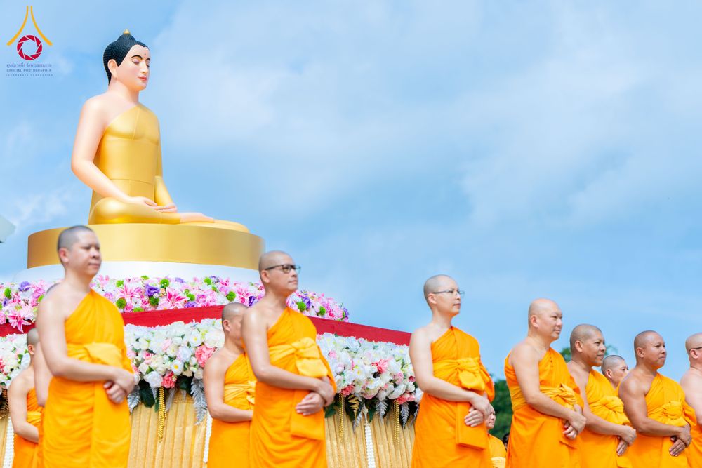คณะสงฆ์ในจีวรสีเหลืองยืนเรียงแถวหน้าพระพุทธรูปสีทองขนาดใหญ่ที่ประดับด้วยดอกไม้