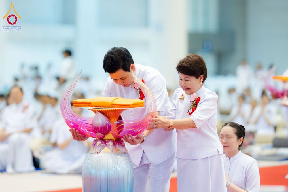 ชายและหญิงสวมชุดสีขาวกำลังประคองพานดอกบัวที่มีผ้าไตรวางอยู่ด้านบนในพิธีทางศาสนา