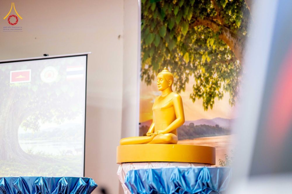พระพุทธรูปสีทองประดิษฐานบนแท่นในห้องประชุมที่มีฉากหลังเป็นภาพต้นโพธิ์และจอโปรเจกเตอร์