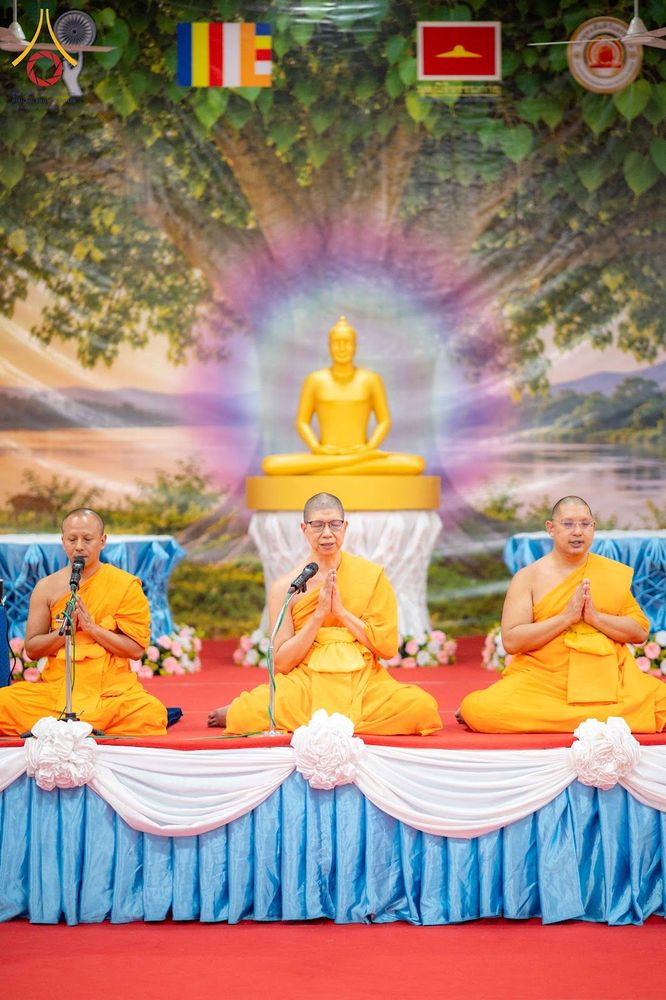 พระสงฆ์สามรูปนั่งพนมมือบนเวทีที่มีฉากหลังเป็นภาพพระพุทธรูปและต้นโพธิ์