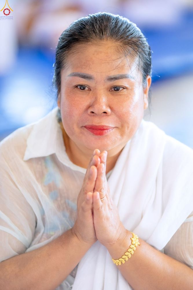 หญิงวัยกลางคนสวมชุดสีขาวกำลังพนมมือไหว้ด้วยท่าทางสำรวมและมีรอยยิ้ม