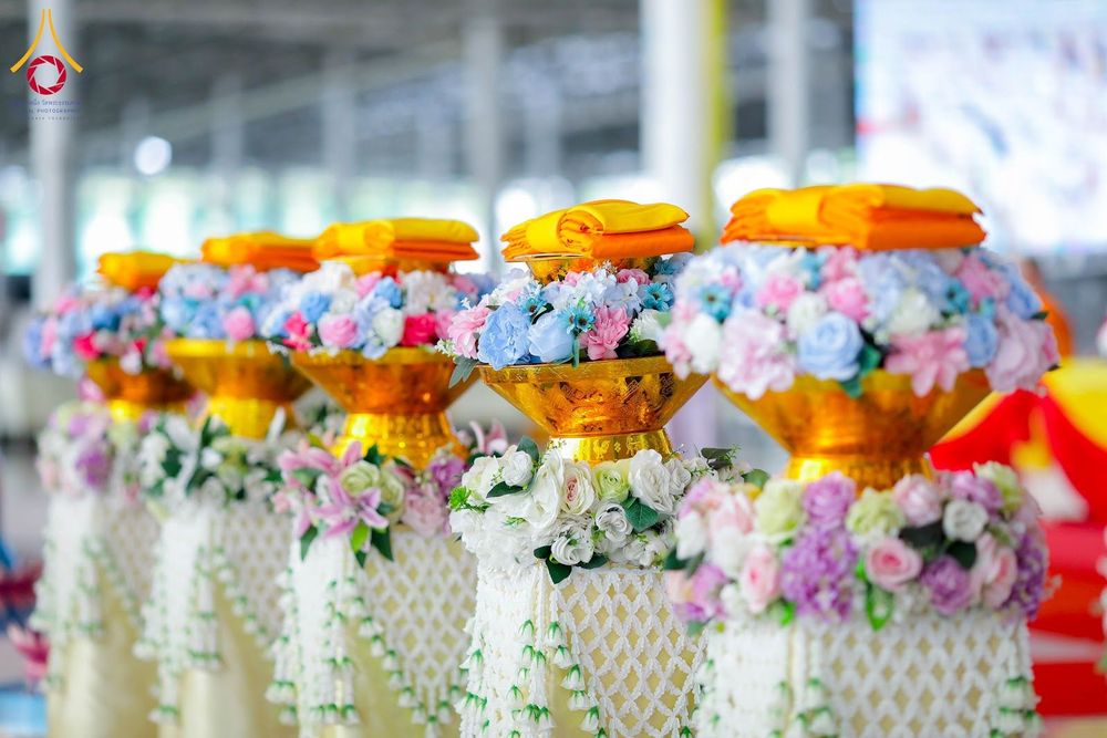 พานพุ่มสีทองประดับดอกไม้และผ้าไตรวางเรียงกันหลายใบในพิธีทางศาสนา