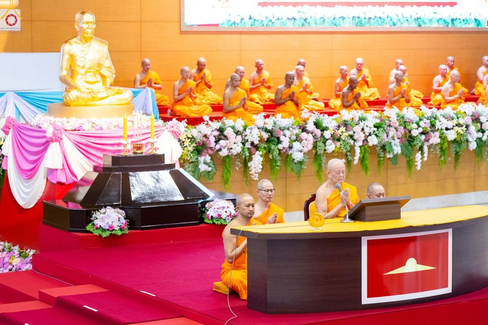 พระภิกษุสงฆ์นั่งประกอบพิธีกรรมทางศาสนาหน้าโต๊ะหมู่บูชาที่มีรูปหล่อทองคำและดอกไม้ประดับในสภาธรรมกายสากล