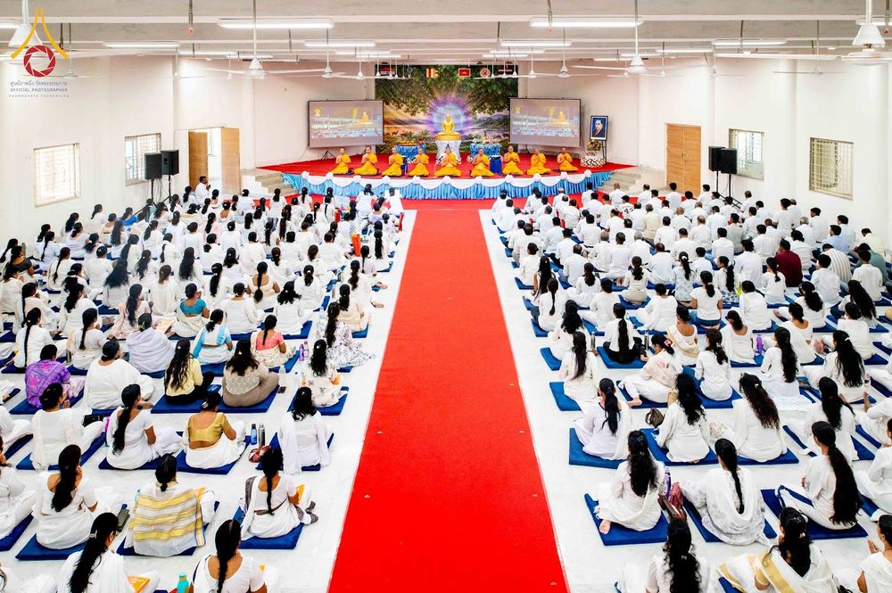 พุทธศาสนิกชนสวมชุดสีขาวนั่งร่วมพิธีในหอประชุมขนาดใหญ่โดยมีพระภิกษุสงฆ์อยู่บนเวทีด้านหน้า