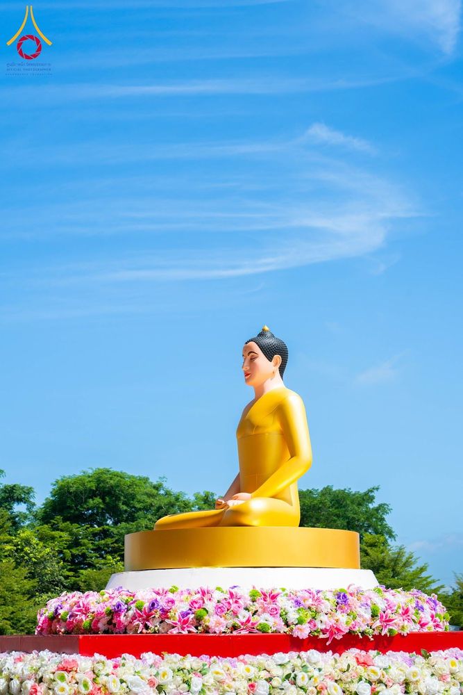 พระพุทธรูปสีทองปางสมาธิประดิษฐานบนฐานที่ประดับด้วยดอกไม้หลากสีสันภายใต้ท้องฟ้าสีคราม