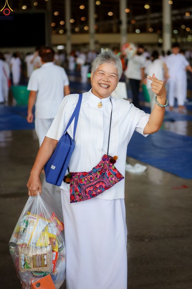 หญิงวัยกลางคนสวมชุดขาวถือถุงอาหารแห้งและทำท่าหัวใจด้วยนิ้วมือ