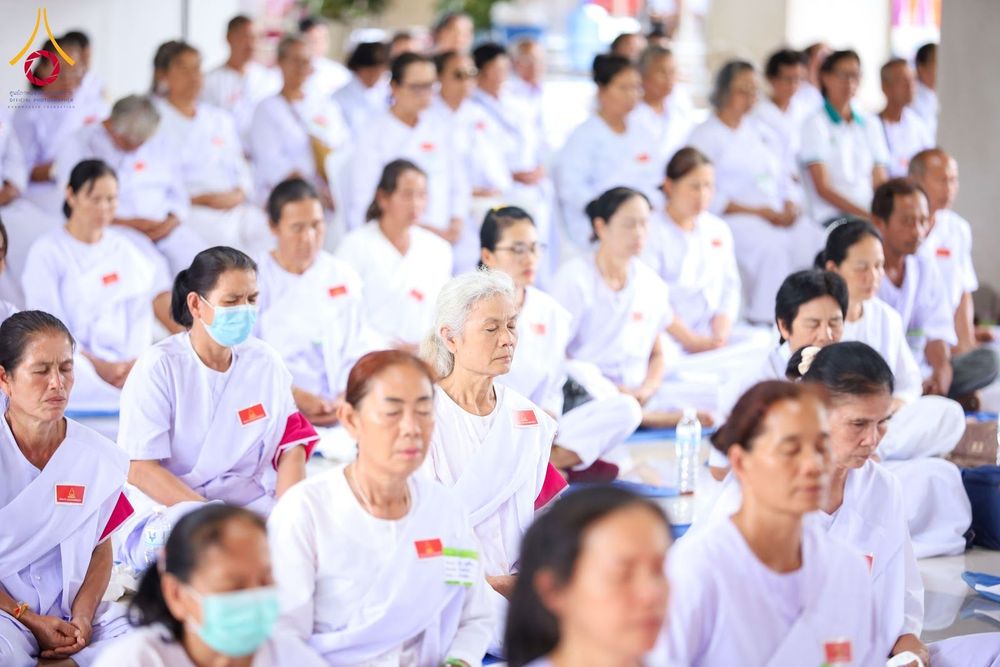กลุ่มคนจำนวนมากสวมชุดสีขาวนั่งสมาธิเรียงแถวกันอย่างเป็นระเบียบภายในอาคาร